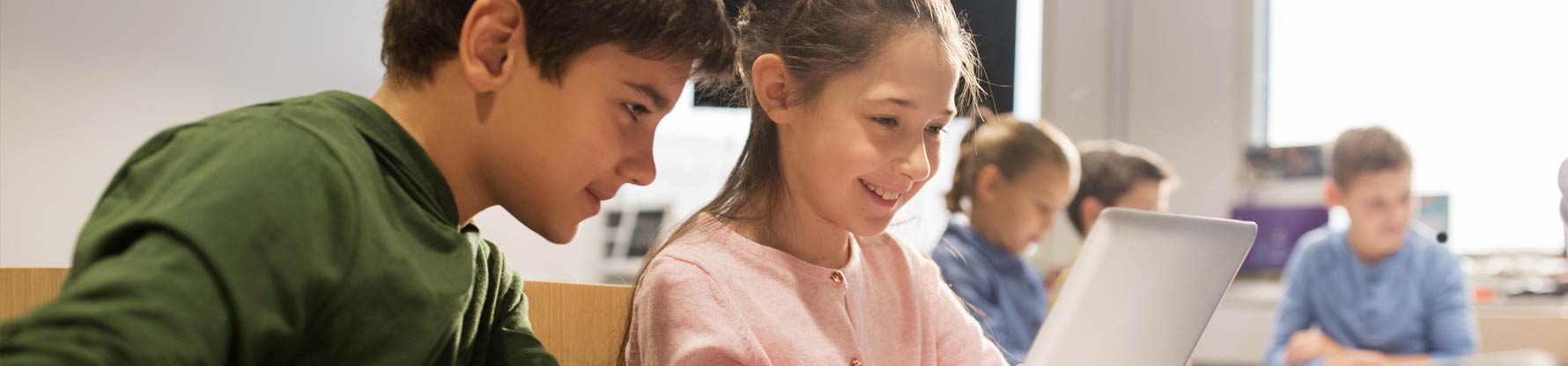 Ein Junge und ein Mädchen sitzen mit anderen Kindern in der Klasse und schauen gemeinsam in einen Laptop